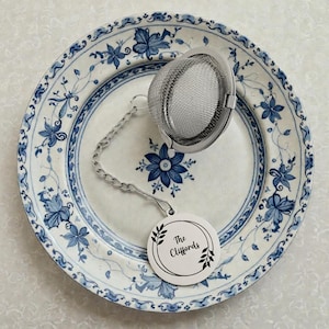May include: A stainless steel tea infuser with a chain and tag that reads "The Cliffords" rests on a white and blue floral patterned plate. The plate has a blue floral design around the rim and center.