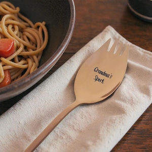 May include: A gold-colored spork with the text "Grandma's Spork" engraved on the head. The spork is resting on a beige napkin next to a bowl of pasta with carrots.