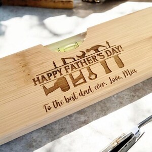 May include: A wooden level with the text "HAPPY FATHER'S DAY!" and various tool illustrations engraved on the surface. The message "To the best dad ever. Love, Mia" is also engraved. The level has a clear vial with a green liquid.
