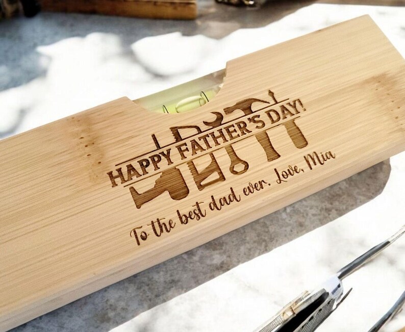 May include: A wooden level with the text "HAPPY FATHER'S DAY!" and various tool illustrations engraved on the surface. The message "To the best dad ever. Love, Mia" is also engraved. The level has a clear vial with a green liquid.
