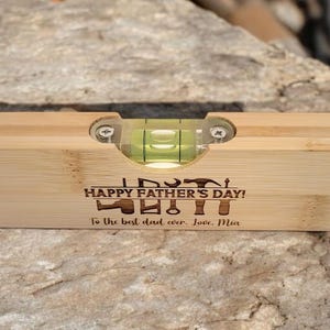 May include: A wooden level with a clear vial containing green liquid. The level has the text "HAPPY FATHER'S DAY!" and "To the best dad ever. Love, Mia" engraved on it. The level is resting on a stone surface.