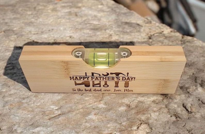 May include: A wooden level with a clear vial containing green liquid. The level has the text "HAPPY FATHER'S DAY!" and "To the best dad ever. Love, Mia" engraved on it. The level is resting on a stone surface.