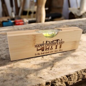 May include: A wooden level with a clear bubble vial. The level is engraved with the text "HAPPY FATHER'S DAY!" and a collection of tools. The inscription reads "To the best dad ever. Love, Mia."