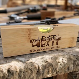 May include: A wooden level with a bubble vial, engraved with "HAPPY FATHER'S DAY!" and tools. The inscription "To the best dad ever. Love, Mia" is also present. The level is made of light-colored wood and rests on a stone surface.