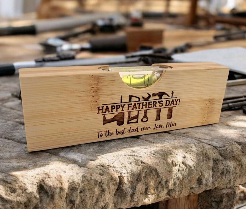May include: A wooden level with a bubble vial, engraved with "HAPPY FATHER'S DAY!" and tools. The inscription "To the best dad ever. Love, Mia" is also present. The level is made of light-colored wood and rests on a stone surface.