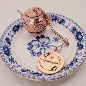 May include: A rose gold tea infuser shaped like a teapot, with a chain and a round tag that says "#1 Grandma". The infuser sits on a white and blue floral patterned plate.