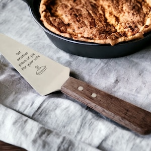 May include: A stainless steel pie server with a wooden handle. The server has the text "Get another piece of pie for your wife" and a pie graphic etched on the blade. A freshly baked pie in a black cast iron pan is in the background.
