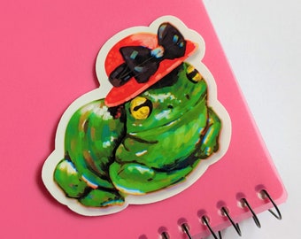 May include: A green frog wearing a red hat with a black bow. The frog is sitting on a white background.