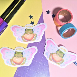 K&ouml;nnte beinhalten: Ein Arrangement mit drei Aufklebern eines Frosches mit Engelsfl&uuml;geln und Heiligenschein. Zwei Stifte mit lila und magenta Tinte und Rollen mit farbigem Washi-Tape sind ebenfalls vorhanden. Der Hintergrund ist in gelbe und rosa Abschnitte unterteilt.