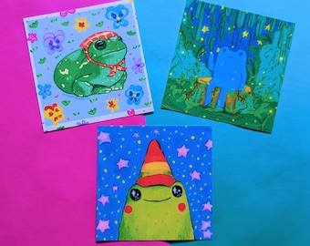 May include: Three illustrated cards featuring frogs. The first card has a green frog wearing a red and white hat and scarf on a blue background with pink and yellow flowers. The second card has a blue frog sitting on a tree stump in a green forest with yellow stars. The third card has a green frog wearing a rainbow hat on a blue background with white stars.