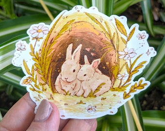 Può includere: Un adesivo con due conigli bianchi che spuntano da una grotta marrone con vegetazione gialla e verde e fiori bianchi. I conigli si guardano e ci sono piccoli cuori rosa sopra di loro.