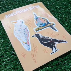 May include: A sheet of stickers featuring three Australian birds: a white cockatoo, a blue and green bird, and a black and white magpie. The stickers are on a light orange background with a pattern of leaves and stars.