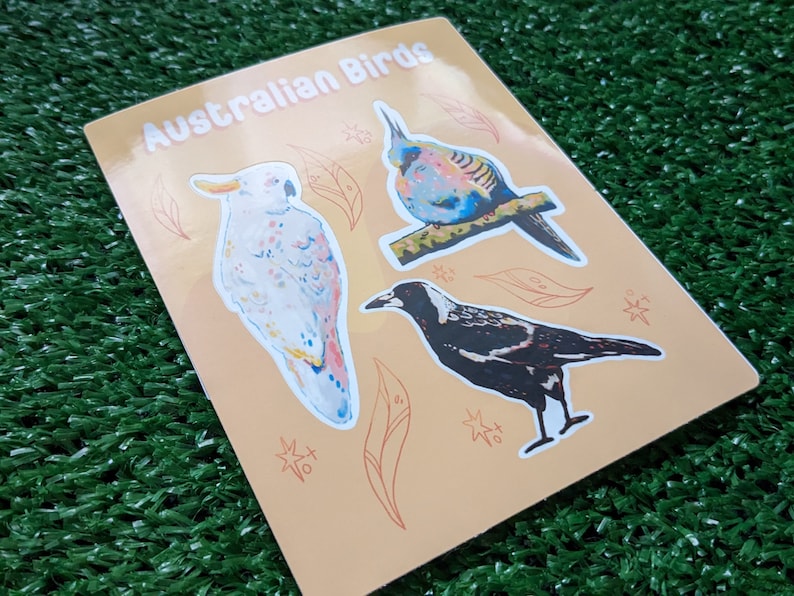 May include: A sheet of stickers featuring three Australian birds: a white cockatoo, a blue and green bird, and a black and white magpie. The stickers are on a light orange background with a pattern of leaves and stars.
