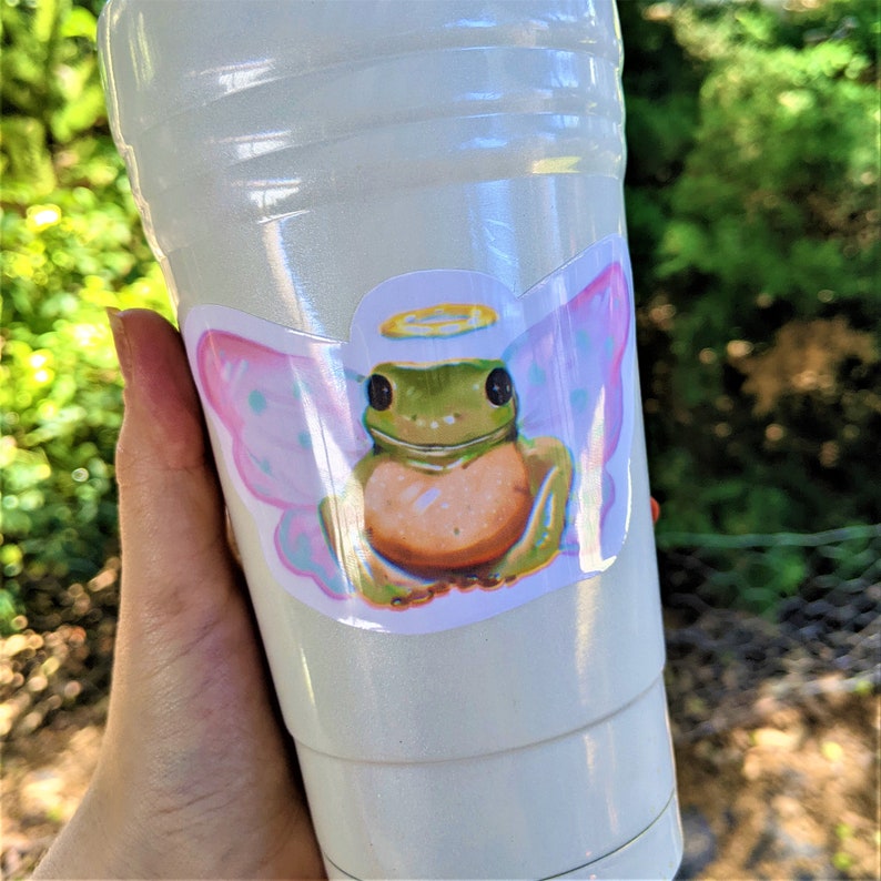 K&ouml;nnte beinhalten: Ein wei&szlig;er Plastikbecher mit einem Aufkleber eines Cartoon-Frosches mit Engelsfl&uuml;geln und einem Heiligenschein. Der Frosch ist gr&uuml;n mit einem orangefarbenen Bauch. Die Fl&uuml;gel sind rosa und blau mit wei&szlig;en Punkten. Der Becher wird von einer Hand gehalten.