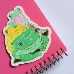 May include: A sticker featuring three frogs stacked on top of each other. The bottom frog is green, the middle frog is yellow, and the top frog is pink. The frogs are on a pink background.