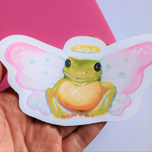 K&ouml;nnte beinhalten: Ein Aufkleber mit einer Cartoon-Fr&ouml;schin mit Feenfl&uuml;geln und einem Heiligenschein. Der Frosch ist gr&uuml;n und gelb, mit gro&szlig;en schwarzen Augen. Die Fl&uuml;gel sind rosa mit hellgr&uuml;nen Punkten. Der Aufkleber wird in einer Hand vor einem rosa-wei&szlig;en Hintergrund gehalten.