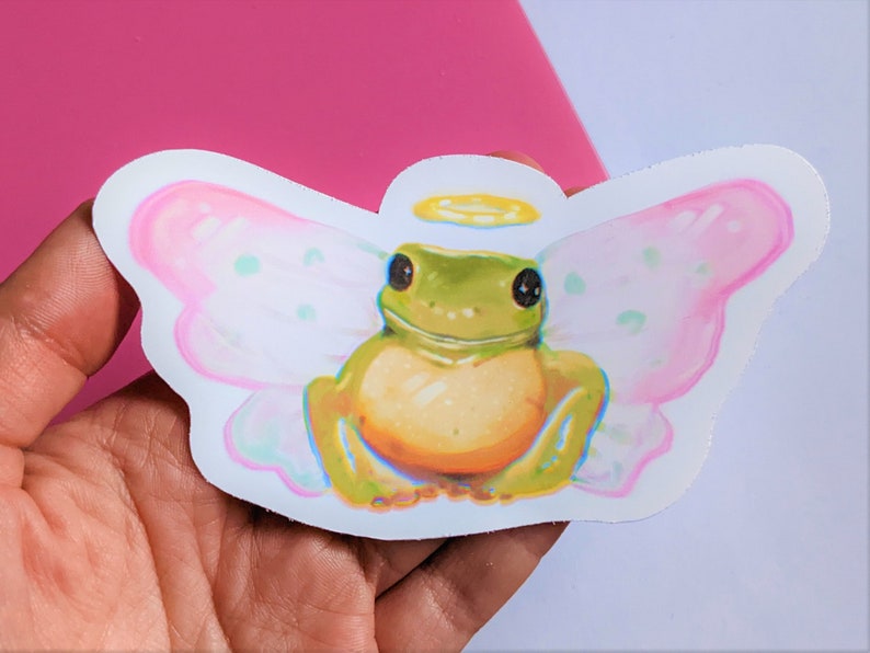 K&ouml;nnte beinhalten: Ein Aufkleber mit einer Cartoon-Fr&ouml;schin mit Feenfl&uuml;geln und einem Heiligenschein. Der Frosch ist gr&uuml;n und gelb, mit gro&szlig;en schwarzen Augen. Die Fl&uuml;gel sind rosa mit hellgr&uuml;nen Punkten. Der Aufkleber wird in einer Hand vor einem rosa-wei&szlig;en Hintergrund gehalten.