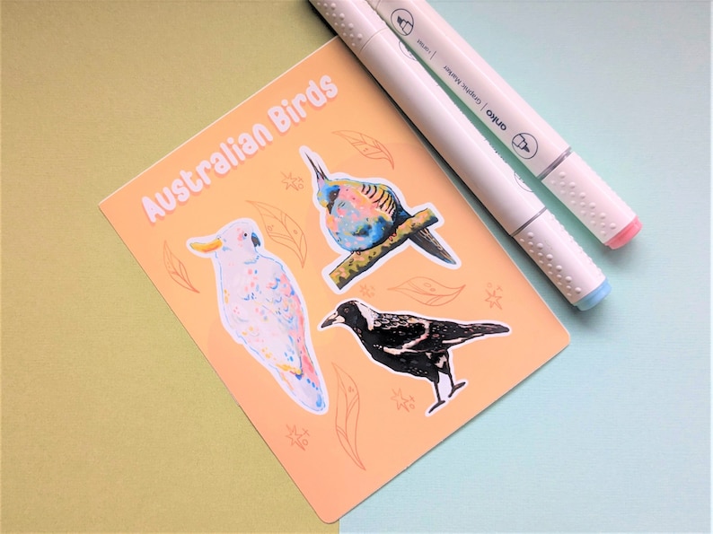 May include: A set of four Australian bird stickers on a peach-colored background. The stickers feature a white cockatoo, a black and white magpie, a blue and green bird, and a brown and white bird. The stickers are surrounded by brown leaves and snowflakes.