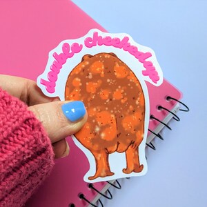 May include: A white sticker with a pink outline featuring a cartoon illustration of a brown, round, furry creature with orange legs and the text "double cheeked up" in pink.