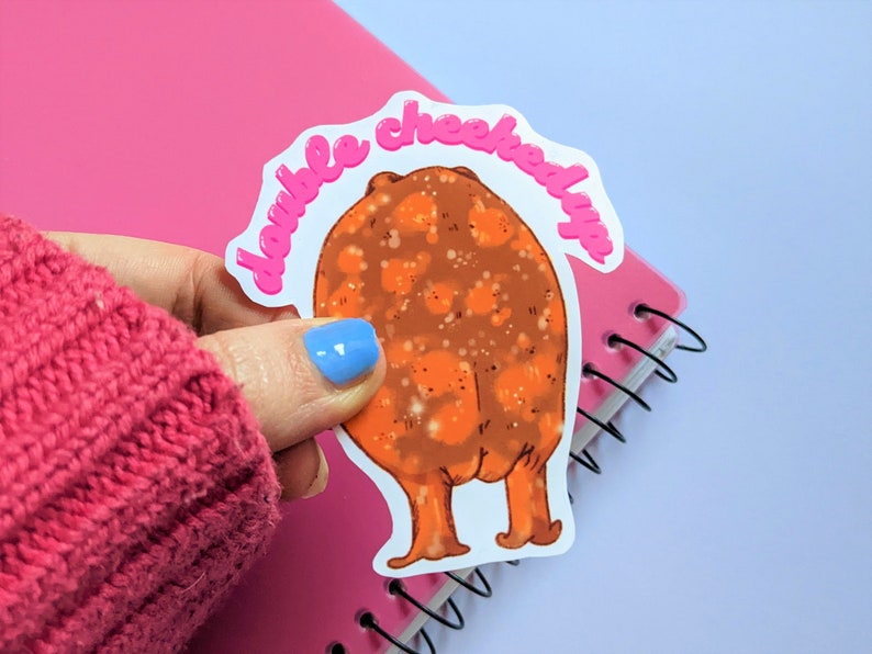 May include: A white sticker with a pink outline featuring a cartoon illustration of a brown, round, furry creature with orange legs and the text "double cheeked up" in pink.