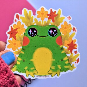 May include: A green frog with yellow and black markings sits in a bed of yellow and orange flowers. The frog has large, sparkling eyes and a small, happy smile. The sticker is signed "eraserheadarts".