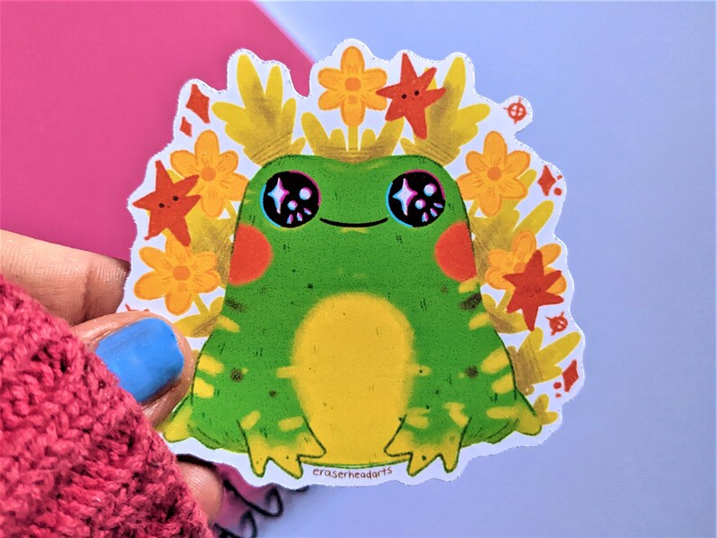 May include: A green frog with yellow and black markings sits in a bed of yellow and orange flowers. The frog has large, sparkling eyes and a small, happy smile. The sticker is signed "eraserheadarts".
