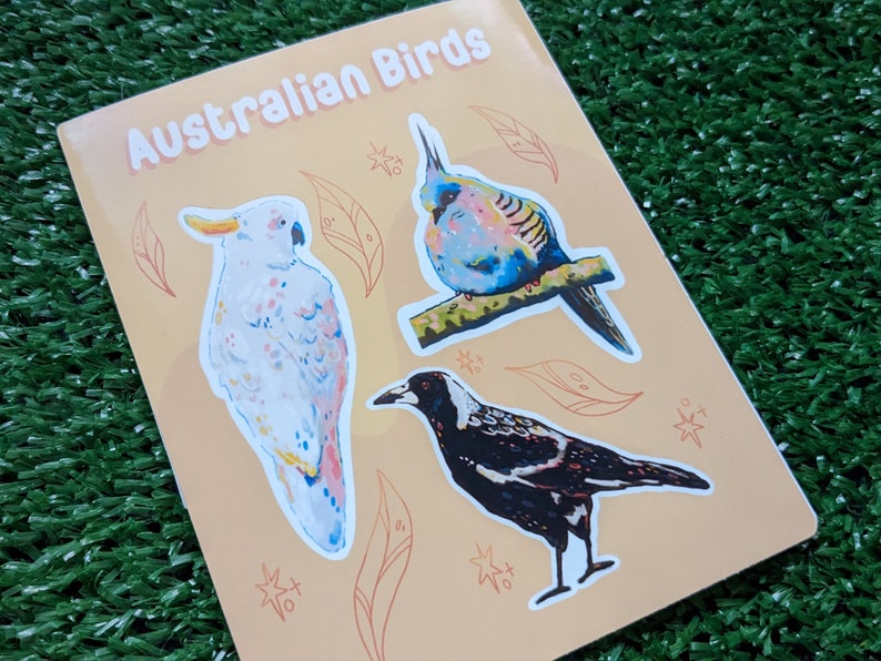 May include: A sheet of stickers featuring illustrations of Australian birds. The stickers are on a light orange background with brown outlines. The birds include a white cockatoo, a blue and green budgie, and a black and white magpie.