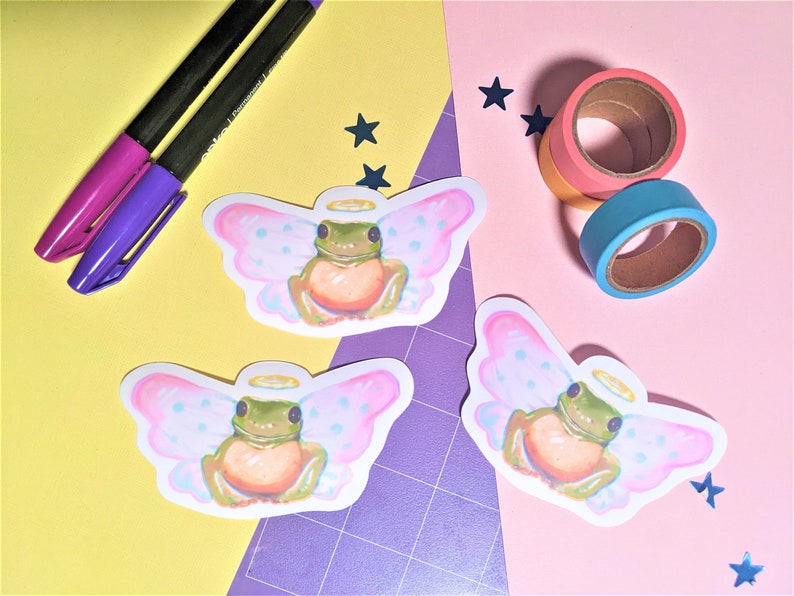K&ouml;nnte beinhalten: Ein Arrangement mit drei Aufklebern eines Frosches mit Engelsfl&uuml;geln und Heiligenschein. Zwei Stifte mit lila und magenta Tinte und Rollen mit farbigem Washi-Tape sind ebenfalls vorhanden. Der Hintergrund ist in gelbe und rosa Abschnitte unterteilt.