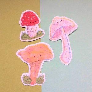 Peut inclure: Trois autocollants de champignons de dessin anim&eacute; mignons avec des visages souriants. Les champignons sont de couleurs et de styles diff&eacute;rents, notamment rouge avec des points blancs, rose avec des points blancs et orange avec des points blancs.