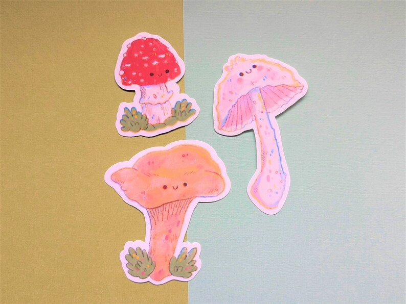 Peut inclure: Trois autocollants de champignons de dessin anim&eacute; mignons avec des visages souriants. Les champignons sont de couleurs et de styles diff&eacute;rents, notamment rouge avec des points blancs, rose avec des points blancs et orange avec des points blancs.