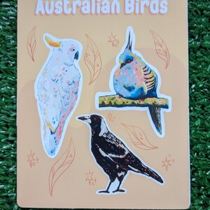 May include: A sticker sheet featuring three Australian birds: a white cockatoo, a blue and pink bird, and a black and white magpie. The sticker sheet has a light orange background with brown leaves and white stars.