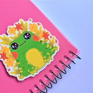 May include: A green frog sticker with big eyes and a yellow belly, surrounded by orange and yellow flowers. The sticker is on a pink notebook with a spiral binding.