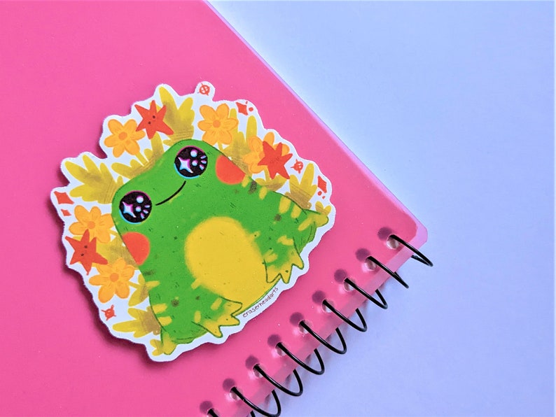 May include: A green frog sticker with big eyes and a yellow belly, surrounded by orange and yellow flowers. The sticker is on a pink notebook with a spiral binding.