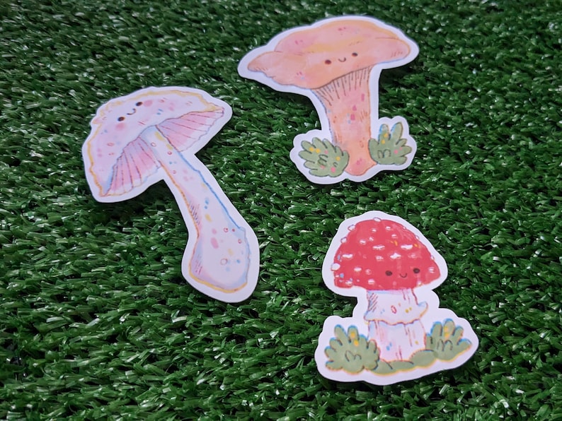 Peut inclure: Trois autocollants de champignons de dessin anim&eacute; avec des visages heureux. Les champignons sont roses avec des points blancs, orange avec des points blancs et rouges avec des points blancs. Les autocollants sont sur un fond vert.