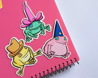 May include: Three frog stickers on a pink notebook. One frog is wearing a pink flower hat, another is wearing a yellow cowboy hat, and the third is wearing a blue wizard hat.