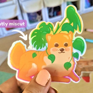 May include: A sticker of a cartoon Shiba Inu dog with a slightly miscut edge. The dog is orange and white and has green leaves around it.