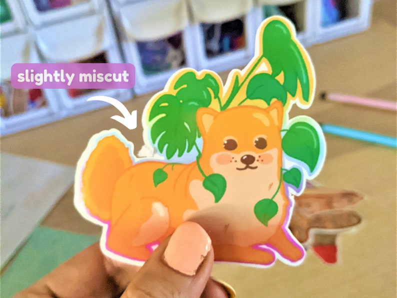 May include: A sticker of a cartoon Shiba Inu dog with a slightly miscut edge. The dog is orange and white and has green leaves around it.