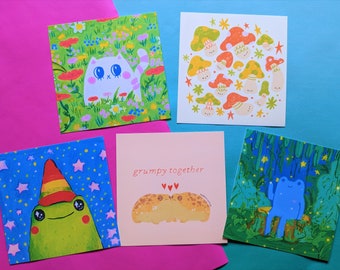 May include: Five square art prints featuring whimsical illustrations. One shows a white cat in a flower field, another depicts mushrooms, and a third has a frog with a party hat. The fourth print says "grumpy together", and the last shows a blue frog in a forest.