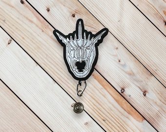 May include: A black and silver metal badge reel with a skull design. The skull is stylized with a pointed top and a large, open mouth. The badge reel is attached to a black cord.