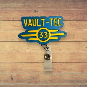 May include: A blue and yellow Vault-Tec badge reel with the text "VAULT-TEC" and the number "33". The badge reel has a retractable cord and a metal clip. The background is a wooden surface.