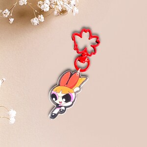 May include: Red and yellow cartoon character keychain with a red flower-shaped clip. The character has black eyes, a pink bow, and a yellow star on her chest.