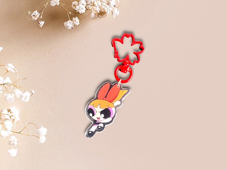 May include: Red and yellow cartoon character keychain with a red flower-shaped clip. The character has black eyes, a pink bow, and a yellow star on her chest.