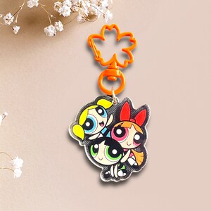 May include: A keychain featuring the Powerpuff Girls, Blossom, Bubbles, and Buttercup, in a cartoon style. The keychain is made of clear acrylic and has a bright orange plastic clip.