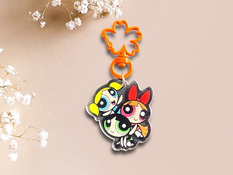 May include: A keychain featuring the Powerpuff Girls, Blossom, Bubbles, and Buttercup, in a cartoon style. The keychain is made of clear acrylic and has a bright orange plastic clip.