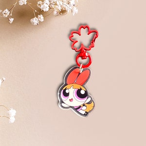 May include: A red and white cartoon character keyring with a red flower-shaped clip. The character has orange hair, pink eyes, and a yellow and black outfit.