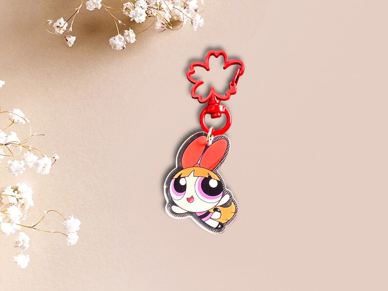 May include: A red and white cartoon character keyring with a red flower-shaped clip. The character has orange hair, pink eyes, and a yellow and black outfit.