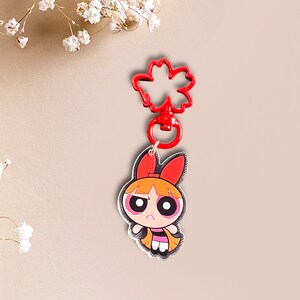 May include: Red and black cartoon character keychain with a red flower-shaped clip. The character has orange hair, red bows, and black eyes.