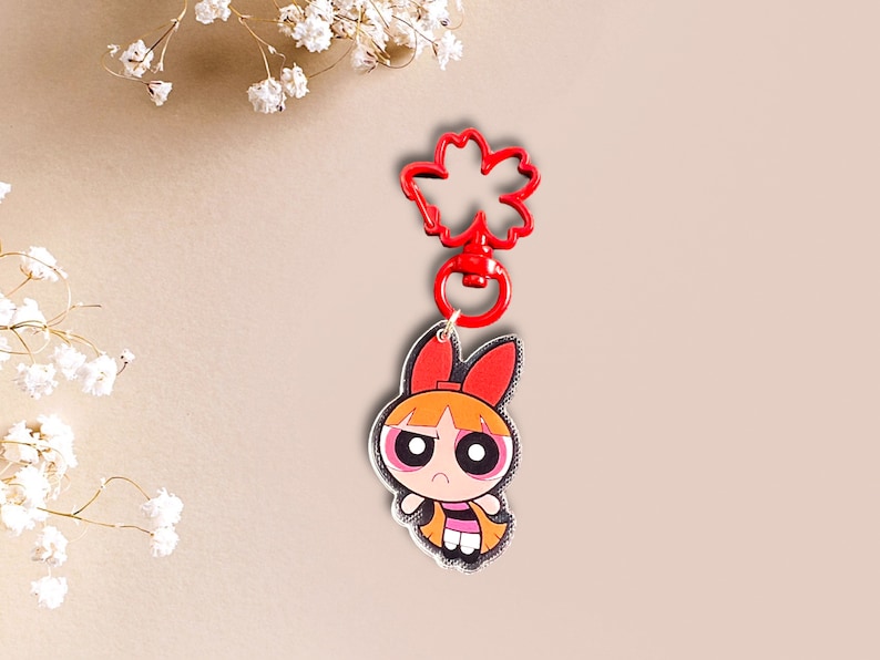 May include: Red and black cartoon character keychain with a red flower-shaped clip. The character has orange hair, red bows, and black eyes.