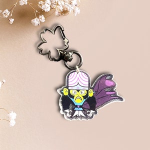 May include: A purple and pink cartoon character with a black cape and a yellow face. The character is attached to a silver keychain with a flower-shaped charm.