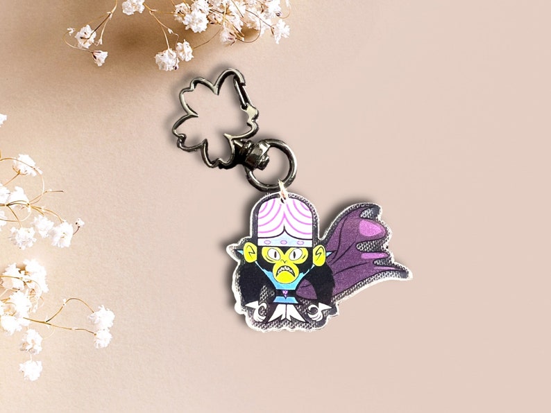 May include: A purple and pink cartoon character with a black cape and a yellow face. The character is attached to a silver keychain with a flower-shaped charm.